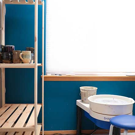 Studio with blue wall and wood shelves with glazed mugs; white pottery wheel and tools.