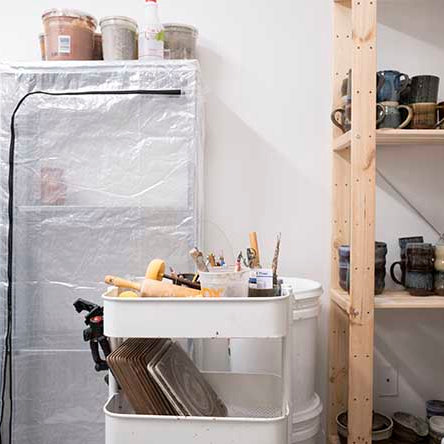 Studio room with shelves featuring handmade mugs and pottery. Some are glazed and some are in progress.